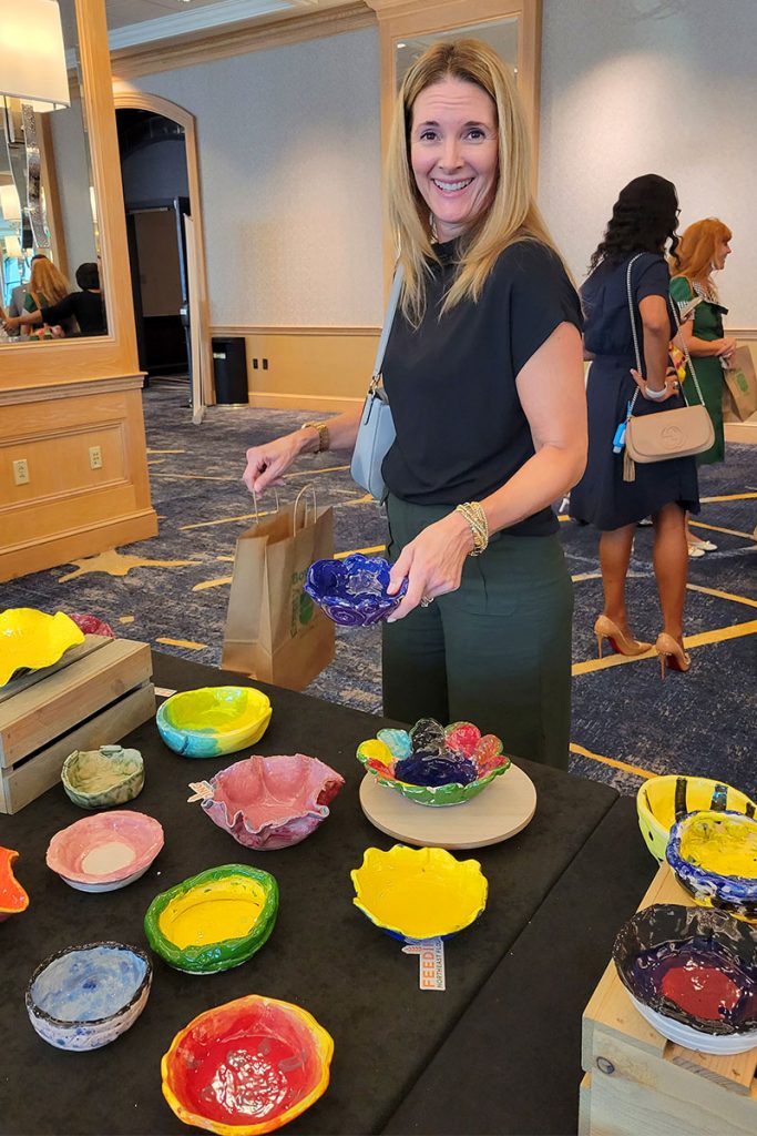 Woman smiling and holding blue bowl in front of table displaying various colorful hand-made bowls