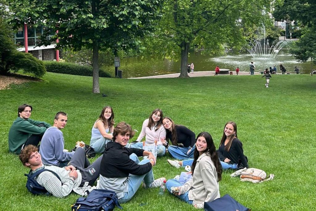 group of students in France sitting on grass with trees and a fountain in the background